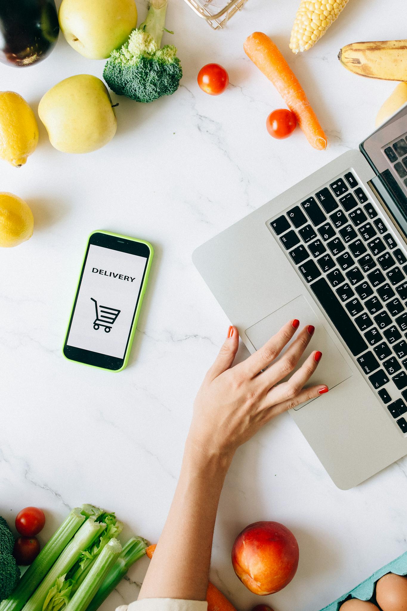 A top view of online grocery shopping using a laptop and smartphone surrounded by fresh vegetables and fruits.