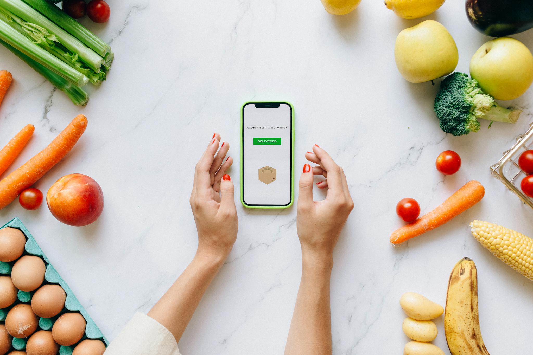 Hands holding smartphone displaying online grocery order amidst fresh vegetables and fruits.