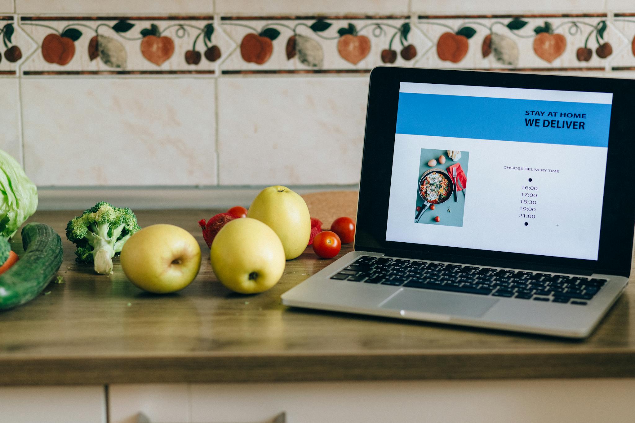 Laptop showing a delivery app beside fresh fruits and vegetables on a kitchen counter.